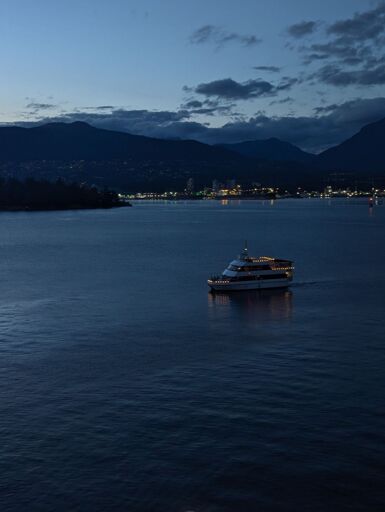 A small boat with 3 floors adorned with lights and populated with partying guests, sailing through an empty waterway at dusk, with mountains and city lights in the distance